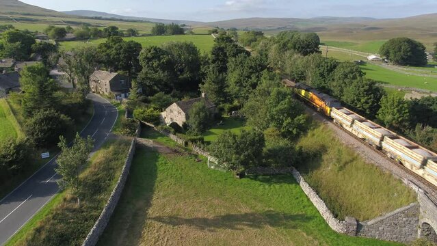 Beautiful summer evening drone footage elevating and slowly panning at the village of Selside, Yorkshire, UK, fields, farmland and rolling hills as a freight train passes under and into the distance