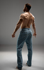 Rear view portrait of strong male model with healthy back posing in denim jeans against grey studio...