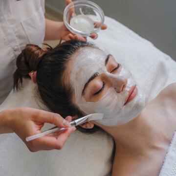 Close up of beautiful young woman lying with closed eyes and cosmetologist applying facial mask by brush in spa salon. People, beauty, spa, cosmetology and