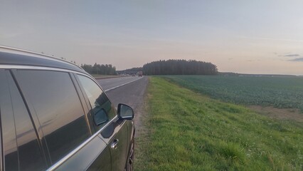 A short stop during a fall road trip. The car is parked on the side of a paved highway with markings and a dividing strip next to a lawn and an agricultural field. Sunset time and light evening haze