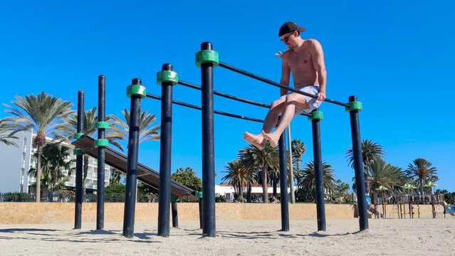 A young male wearing shorts, a baseball hat and sunglasses doing triceps dips on parallel bars at an outdoor gym on the beach