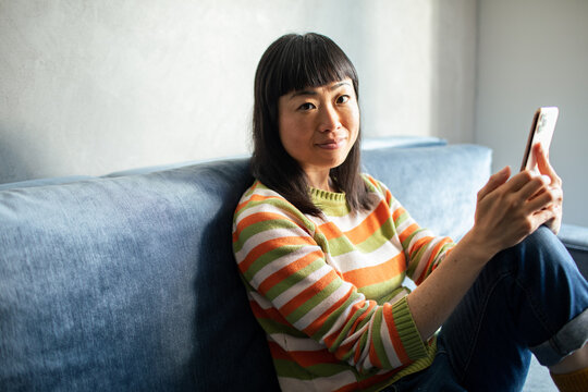 Asian young woman using smartphone on sofa looking at camera