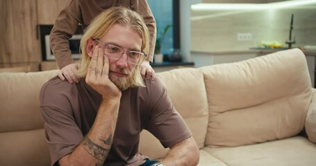 A tortured blond man with a beard and glasses sits on the sofa while his little albino son pesters him and wants to play with the man at home in a modern apartment. Tired man with glasses cannot pay