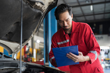 Male mechanic holding clipboard during working and checking car engine under hood of car in auto...