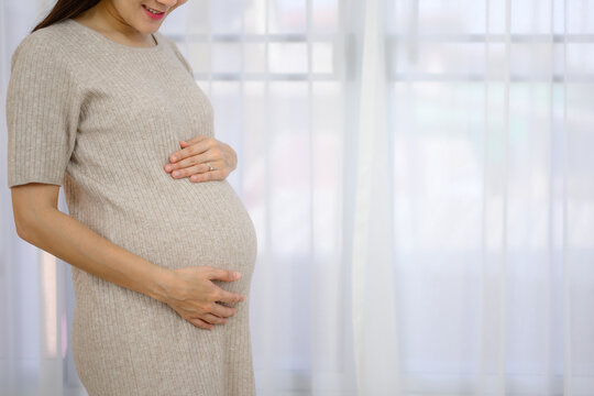 Picture of pregnant woman with pregnancy holding her large belly standing against the window in a white room at home.