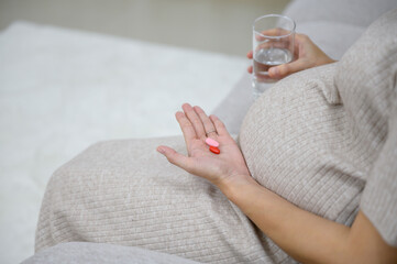 Close-up photo of a pregnant woman taking pregnancy medicine. Pregnant woman holds pills and glass of water in her home living room with prenatal care - obrazy, fototapety, plakaty