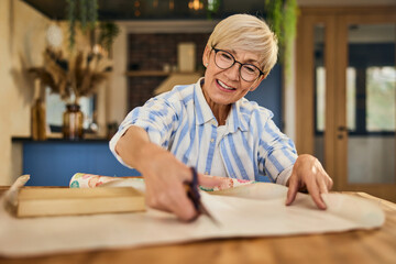 A smiling mature female, using scissors and cutting wrapping paper, packing a book for a gift.
