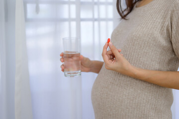 An image of a pregnant young woman takes pregnancy medicine Pregnant woman holds pills and glass of water in her home living room with prenatal care