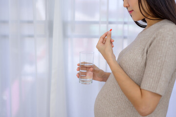Asian pregnant woman takes pregnancy medicine as prescribed by her doctor. Pregnant woman holds pills and glass of water in her home living room with prenatal care