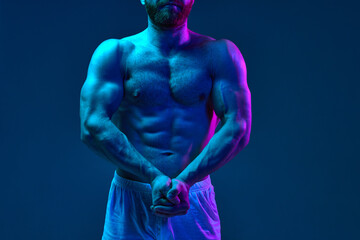 Shirtless fit male model with crossed arms posing gym shorts in neon lighting against blue studio background. Concept of beauty and fashion, art, aesthetic of body, masculinity. Ad.