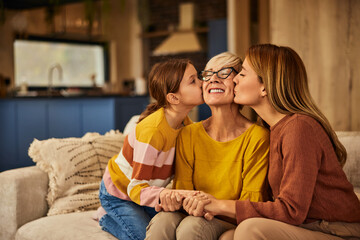 Happy grandma getting kisses from her daughter and grandchild, sitting on the sofa.