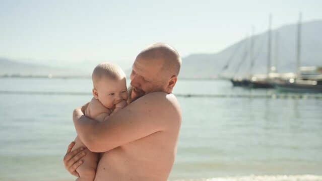 Authentic shot of young father caressing and kissing his baby standing by the sea. Dad and little boy hug during summer vacation. Concept of family, fatherhood, motherhood, parenthood, childhood, life