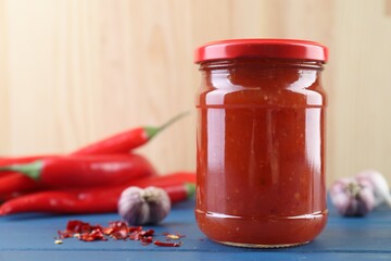 Spicy chili sauce in jar, garlic and peppers on blue wooden table, space for text