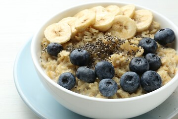 Tasty oatmeal with banana, blueberries and chia seeds served in bowl on white wooden table, closeup