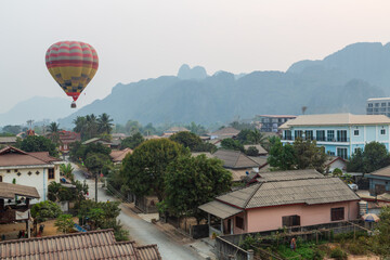 VANG VIENG, LAOS - February 2020: Hot air balloons over the street with local houses and a road. Misty hills in the background