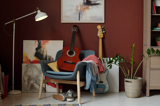 Soft comfortable armchair with acoustic guitar and pile of clothes standing by wall with paintings, lamp, green plant and electric string instrument