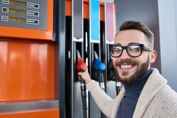 Portrait of modern man dressed in casual clothes stopping at gas station to refuel car, picking up nozzle from pump and smiling looking at camera - obrazy, fototapety, plakaty