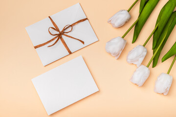 White envelope and blank card on a peach background with white tulips. Congratulations concept for Easter, International Women's Day, Mother's Day, Valentine's Day and Birthday.