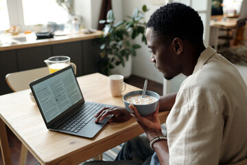 Young African American businessman with bowl of cornflakes sitting in front of laptop screen during...
