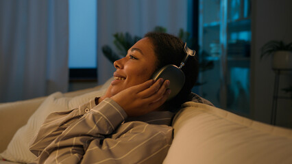 Relaxed African American woman in headphones fall on comfortable cozy sofa evening night leisure at home listening music ethnic girl rest with eyes closed enjoy sound in earphones sing relaxing song