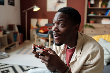 Young amazed African American man with gamepad sitting in front of tv set or computer screen and...