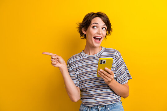 Photo of impressed funny woman dressed t-shirt holding smartphone directing staring at empty space isolated on yellow color background