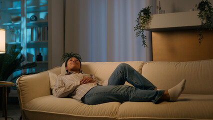 Tired African American woman in evening night living room lying on cozy sofa falling down on...