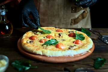 The chef puts spinach on a pizza with mushrooms, tomatoes and cheese. Pizzaiolo in a leather apron prepares pizza.