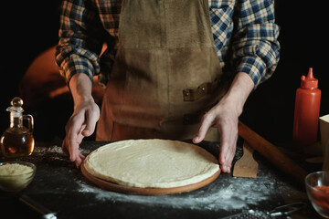 The chef rolls out the dough with a rolling pin. A guy in a leather apron is preparing pizza....