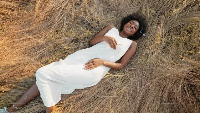 Beautiful black woman with afro hair in white dress lies on grass smiles rejoices slide turn in freedom holds an ear of wheat above her on walk in summer at sunset slide turn. Go Everywhere lifestyle