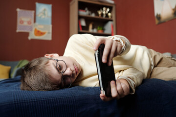 Youthful pre-teen boy in eyeglasses and casualwear relaxing on bed and looking at mobile phone...