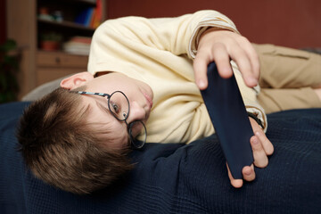 Restful schoolchild looking at smartphone screen while lying on bed in his bedroom and messaging with someone or watching online video
