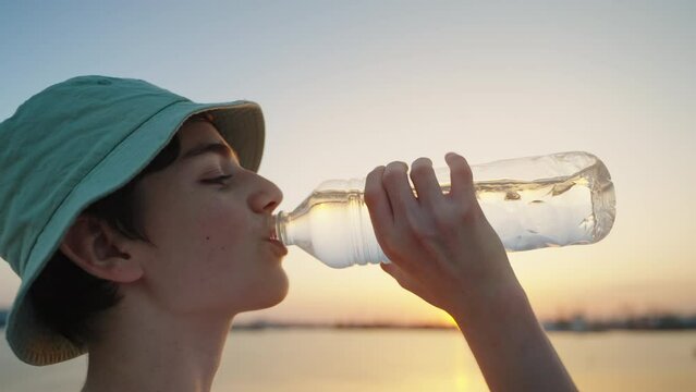 Male drinks water from bottle greedily in heat after cycling on walk summer vacation after exercises or sport sunset. Boy caucasian drinking mineral water on hot day in cap.  Lifestyle. Abnormal heat