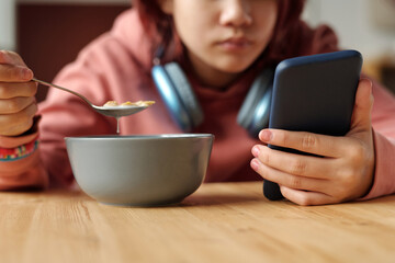 Hand of teenage girl with mobile phone having cornflakes with milk from bowl while sitting by table...