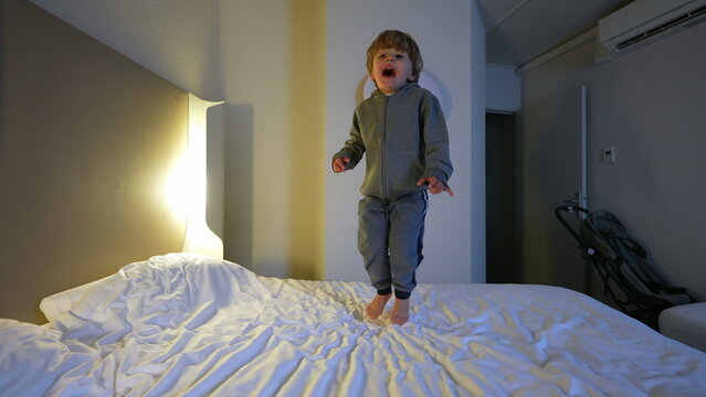 Happy young 3 year old boy bouncing in hotel bed, carefree excited child having fun while traveling jumping up and down on bedsheets