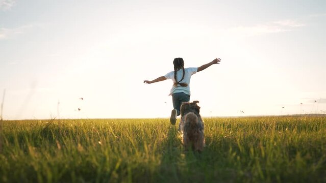Cheerful child run along green grass. Dog run after girl along green glade in summer. Child play with dog in park. Happy girl walk in summer in park with dog. Child in rays of sun in field on grass.