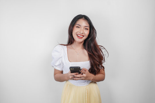A cheerful Asian woman is smiling and holding her smartphone, isolated by white background.