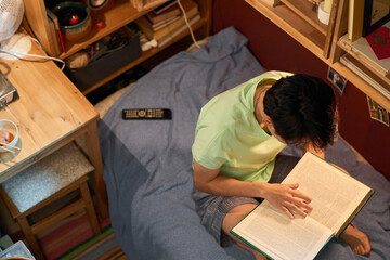Above angle of young Chinese female student sitting on bed in small apartment and reading encyclopaedia while preparing homework