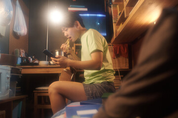 Modern Chinese girl with mobile phone watching online video or communicating in chat while eating wok in small apartment