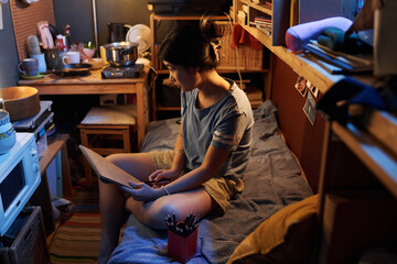Young Asian female student sitting on bed in small apartment and drawing kitchenware with pencil in...