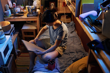 Young student with pencil and notepad sitting on bed in small apartment and drawing kitchenware standing on top of microwave oven