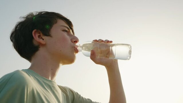 Male teenager drinks water from bottle greedily in heat. Abnormal heat after cycling on walk summer vacation after exercises or sport sunset. Boy caucasian drinking mineral water on hot day. Lifestyle