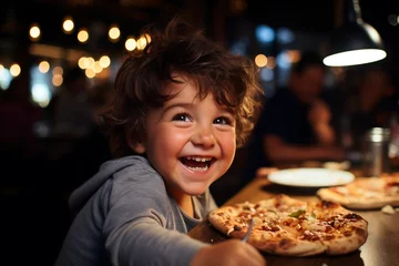 Foto op Plexiglas A happy laughing curly-haired boy at the table is eating fresh pizza, holding food in his hands. A joyful atmosphere in the pizzeria © Irina Afanaseva