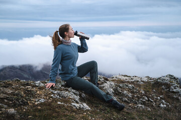 A young beautiful girl with straight brown hair in a ponytail drinks from a thermal glass in nature, tourist resting outdoors, in full view