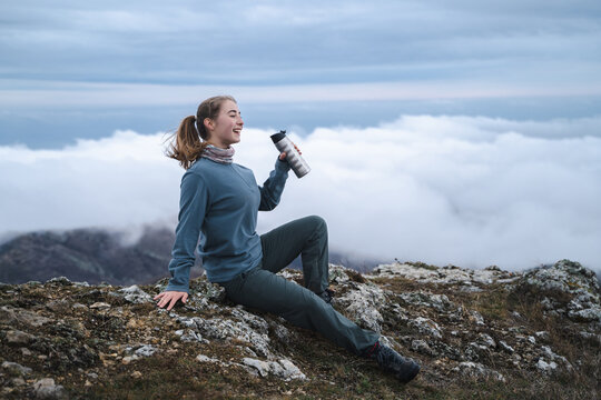 Young woman taking a break to drink from water bottle while hiking, hiker on top of a mountain among the clouds, a tourist enjoys the views at a rest stop