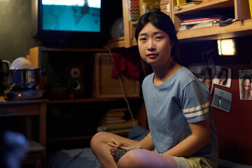 Young Asian woman with chopsticks and empty bowl in hands sitting on bed in small apartment and looking at camera after lunch