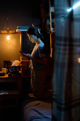 Young woman with cellphone watching online video or texting while standing by electric stove in the kitchen of microflat and cooking