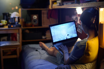 Rear view of young woman sitting on bed and pressing buttons of laptop keyboard while watching...