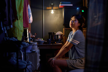 Young Chinese woman in casualwear sitting on bed in small apartment and looking at raincoats or jackets hanging on rope along wall