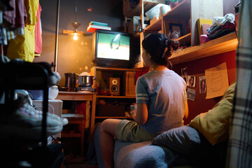 Rear view of young Asian woman in grey t-shirt sitting on bed in front of screen of TV set and...